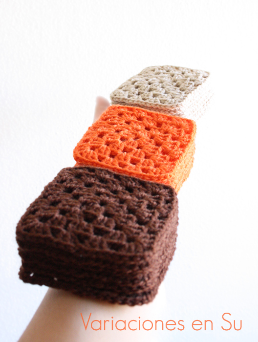 Brazo sujetando pilas de granny squares o cuadrados tejidos a ganchillo en lana de colores marrÃ³n, naranja y beige.